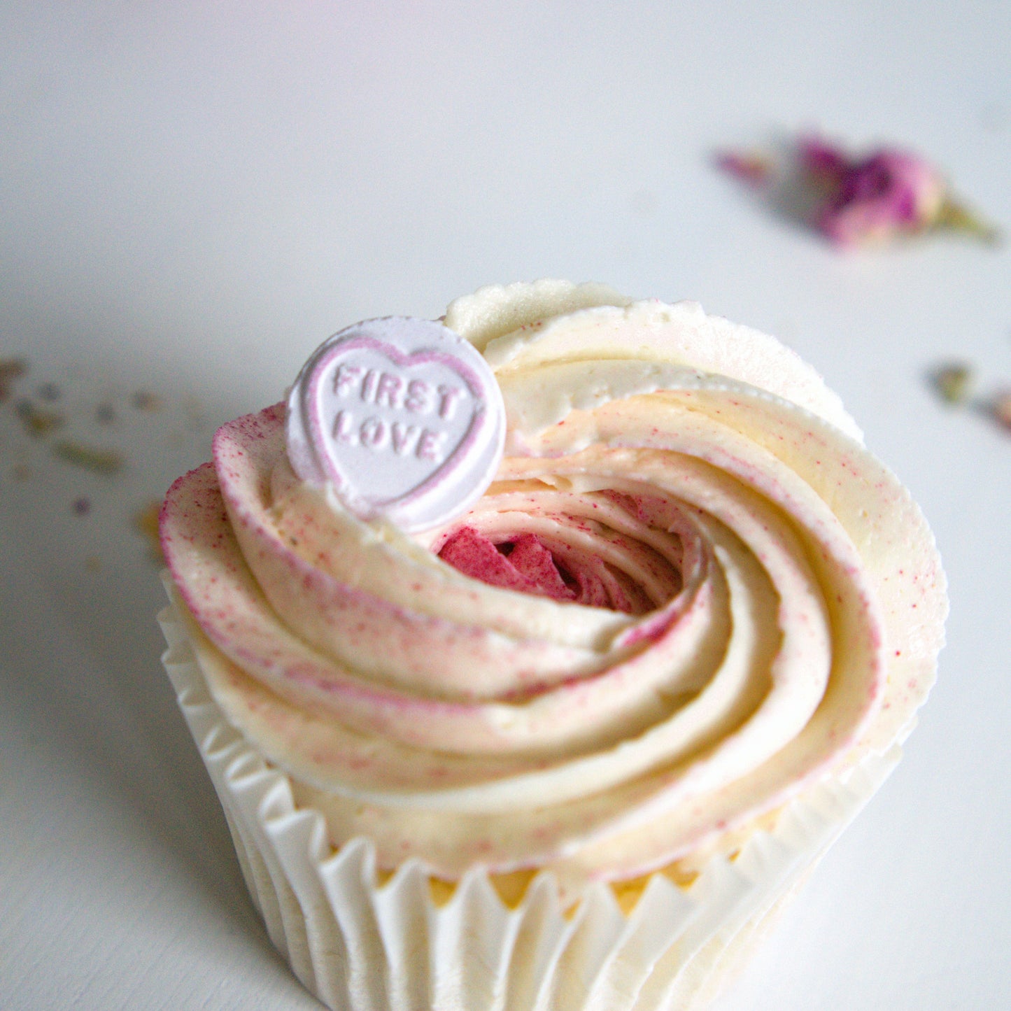 Cupcake with pink and white frosting and a heart-shaped 'First Love' topper on a light background.