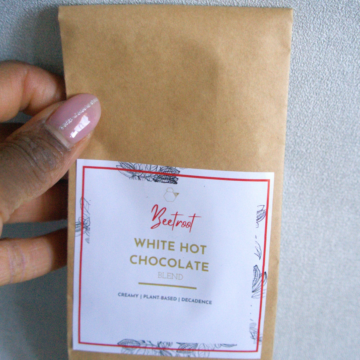 Kraft paper package of beetroot white hot chocolate held by a hand against a light gray background