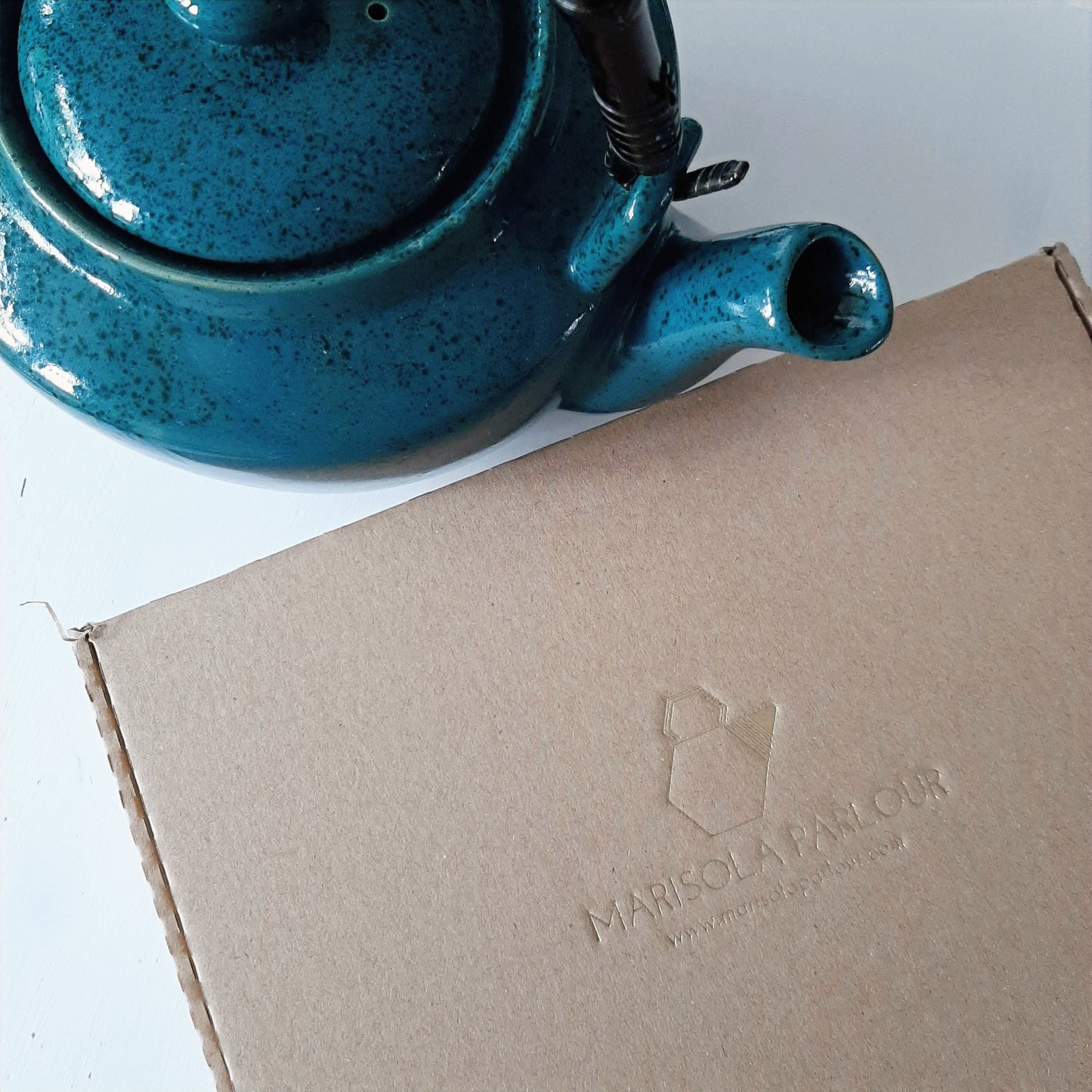 Blue ceramic teapot next to a cardboard box with 'Marisola Parlour' branding.