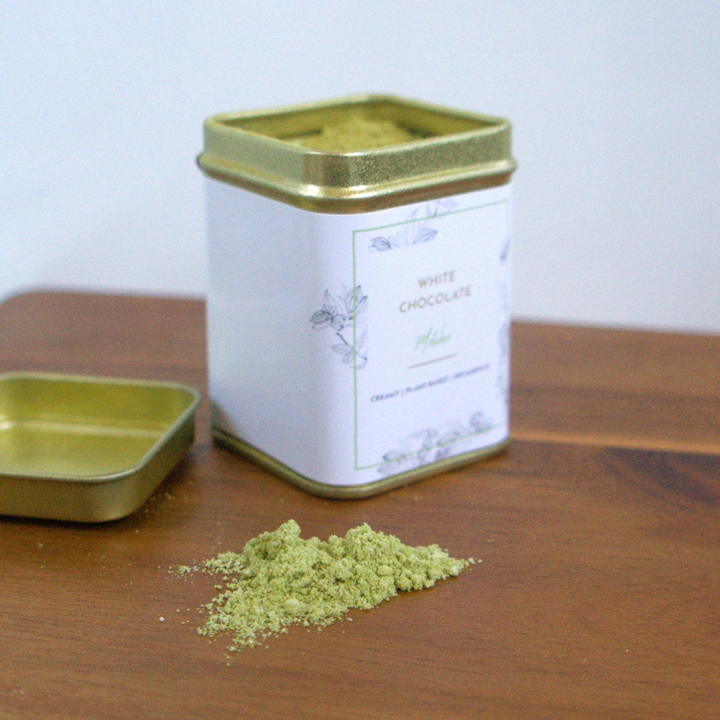 A gold tin with a white label and green matcha powder on a wooden surface.