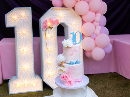 Two-tier tenth birthday cake displayed beside illuminated number 10 decorations and a pink balloon backdrop.
