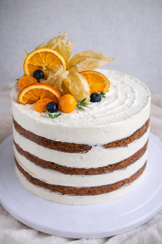 Naked gluten-free and vegan carrot cake with white frosting decorated with orange slices, blueberries, and dried fruit on a white background.
