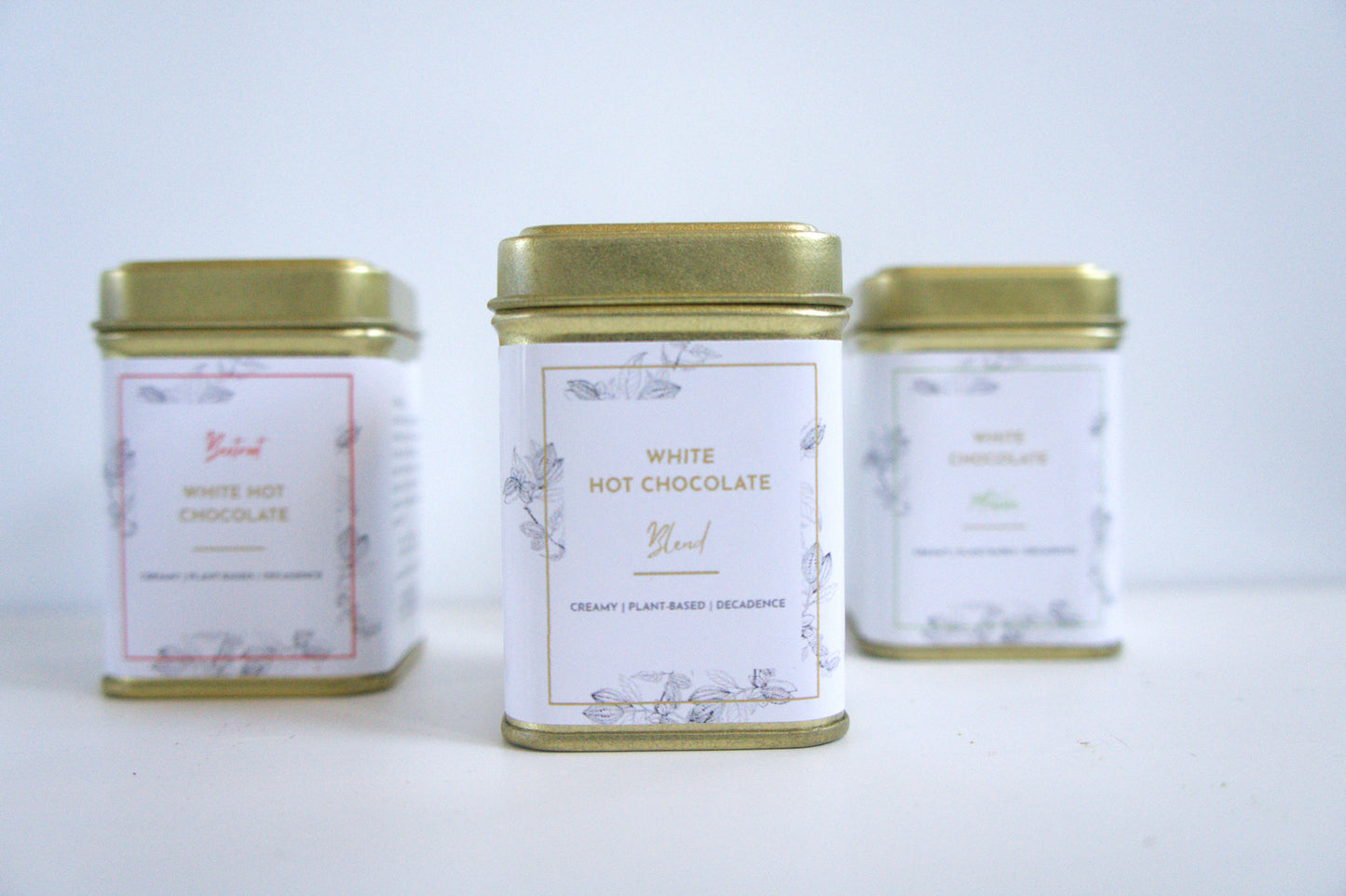 Three tins of vegan white hot chocolate on a light grey background