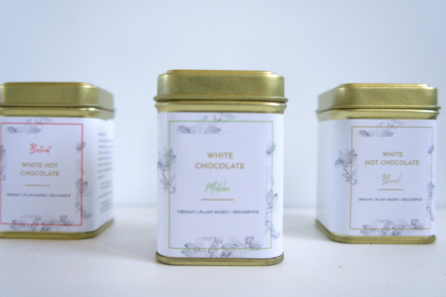 Three tins of vegan white hot chocolate with gold lids on a light grey background