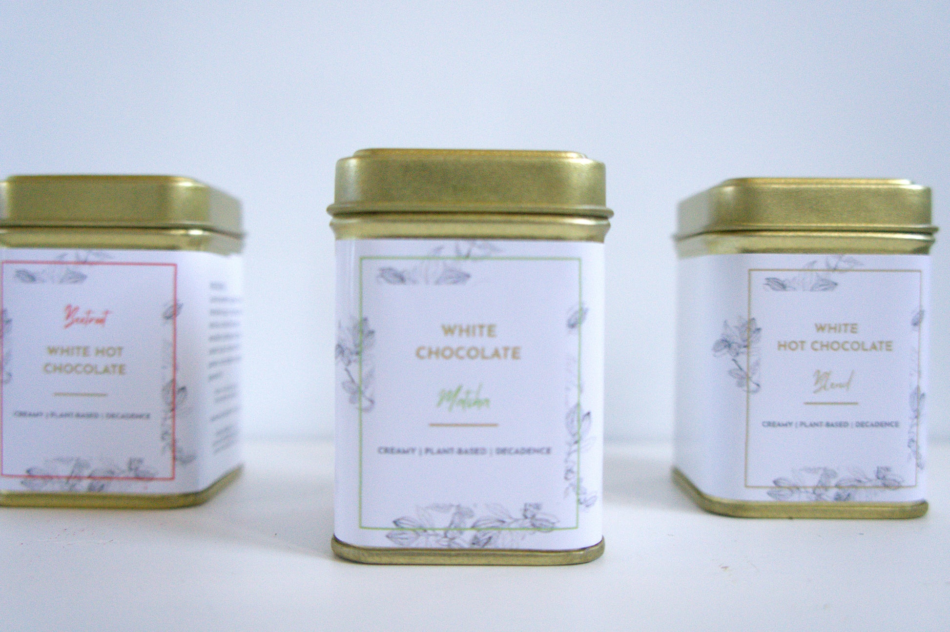 Three tins of vegan white hot chocolate with gold lids on a light grey background