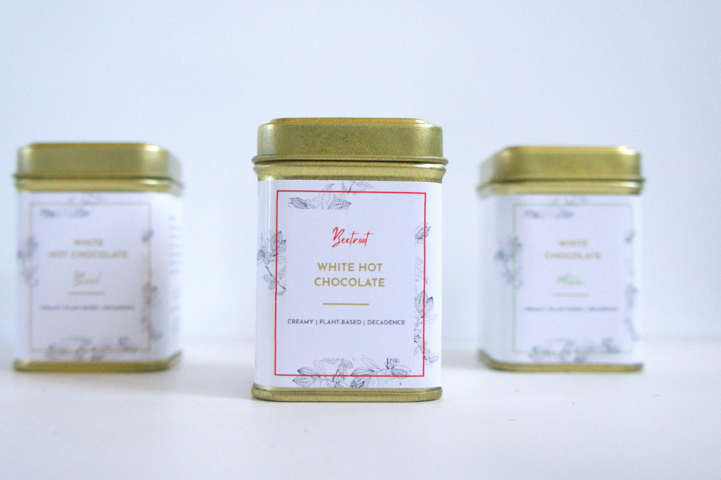 Three tins of vegan white hot chocolate with gold lids on a light grey background