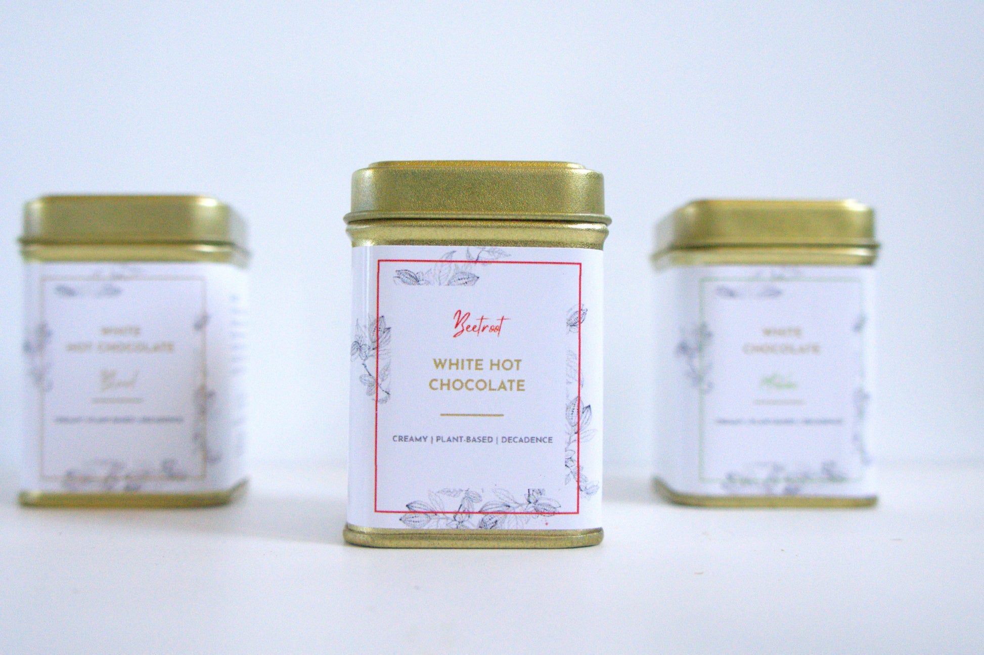 Three tins of vegan white hot chocolate with gold lids on a light grey background