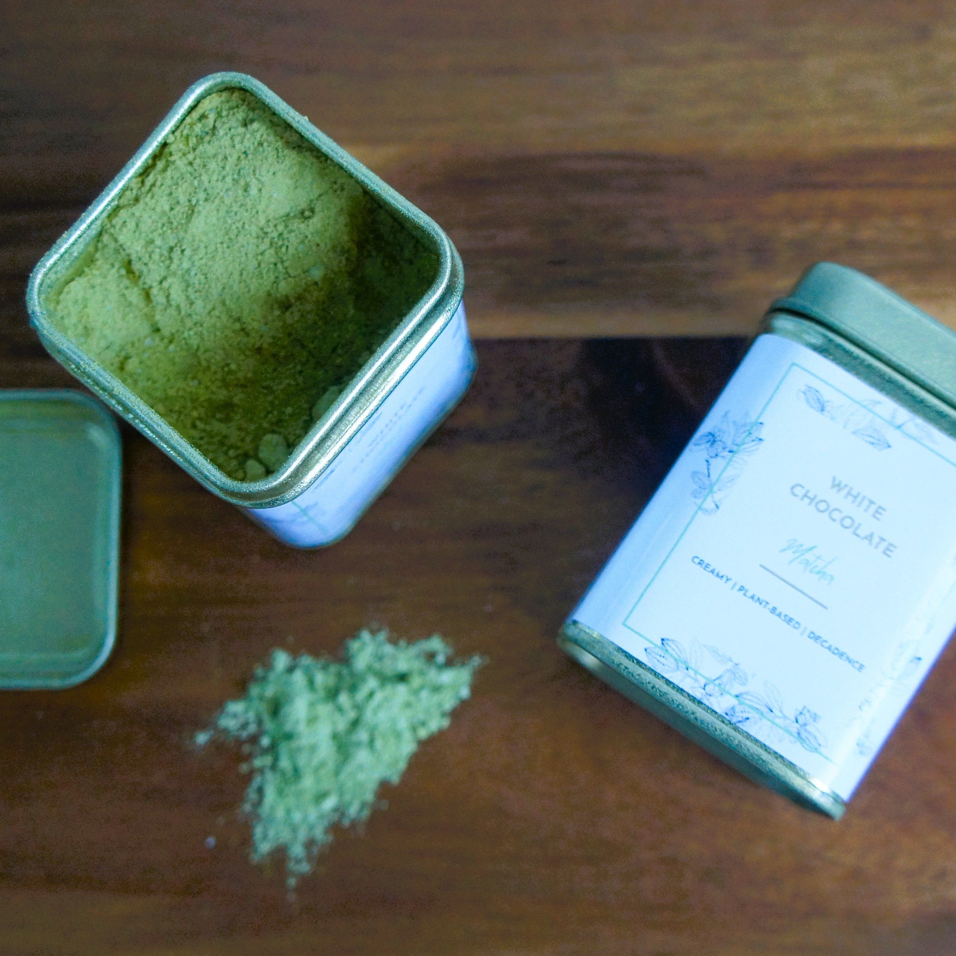 Two gold tins on a wooden surface with one open showing green matcha powder.