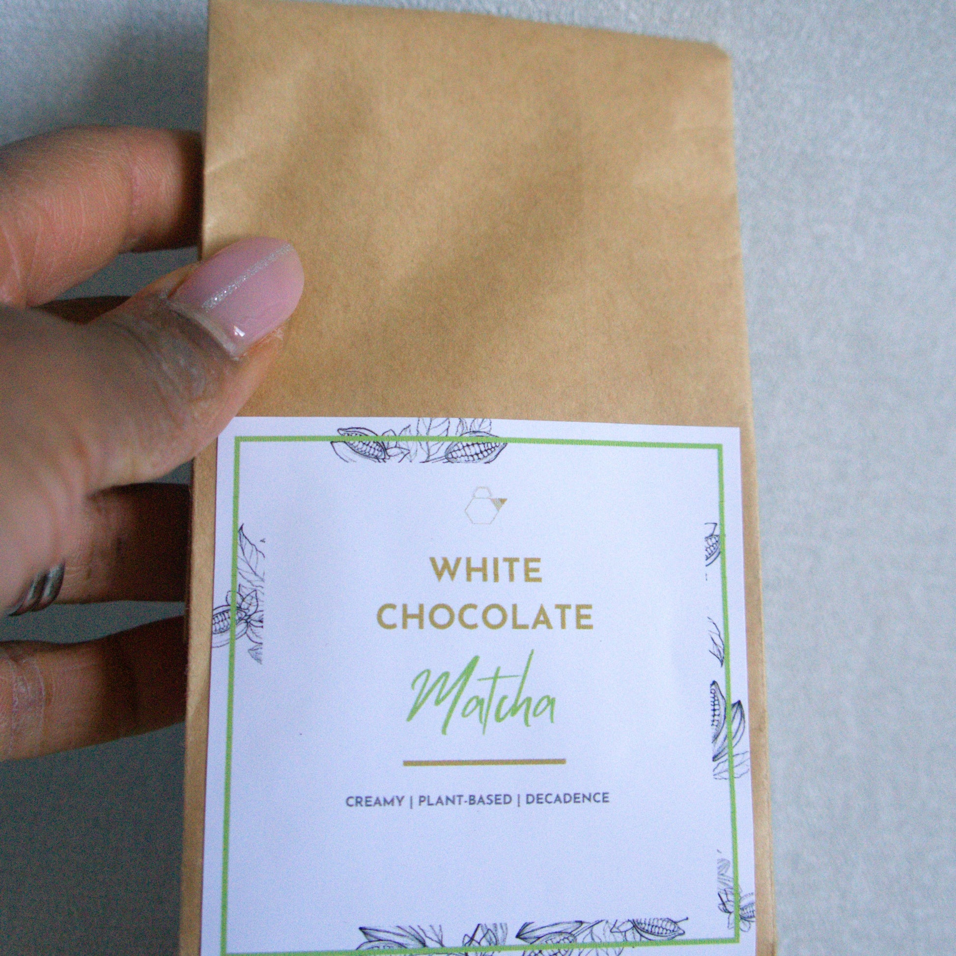 Kraft paper packaging with a white label featuring 'White Chocolate Matcha' text, held by a hand against a light gray background.