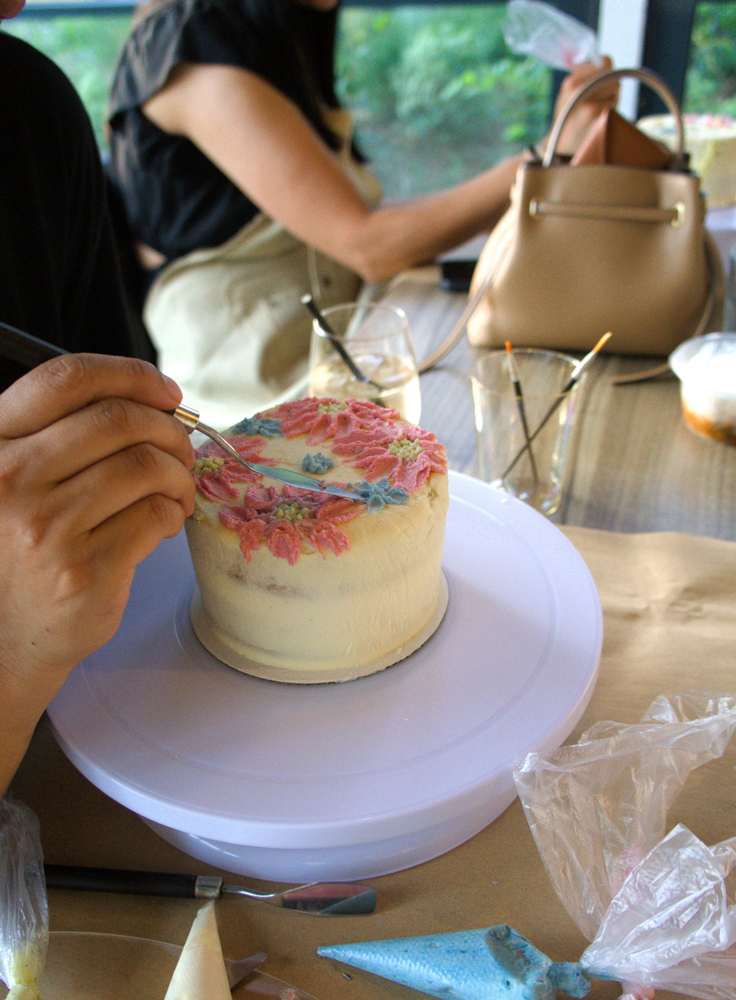 A close-up of a person's hand painting a floral design on a frosted cake sitting on a white rotating display stand. The scene is set in a bakery or studio with other tools, a beige handbag, and another person blurred in the background.