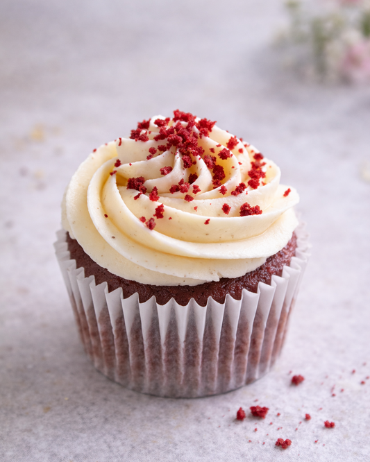 Red velvet cupcake with cream cheese frosting and red sprinkles on a light surface.