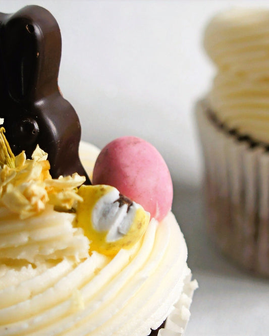 Close-up of a cupcake with white frosting, chocolate bunny, pink Easter egg, and yellow flowers on a white background.
