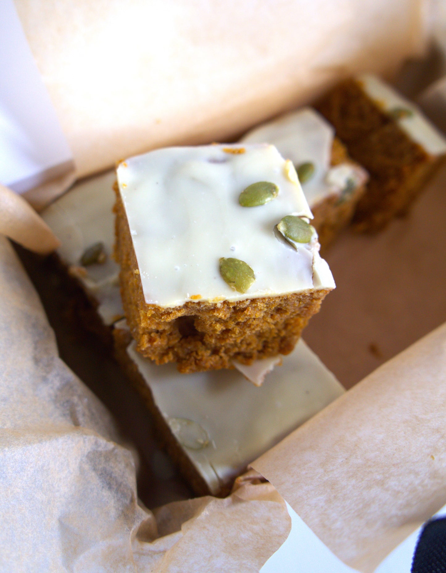 Square piece of blondie cake with white frosting and green pumpkinseeds in a box.