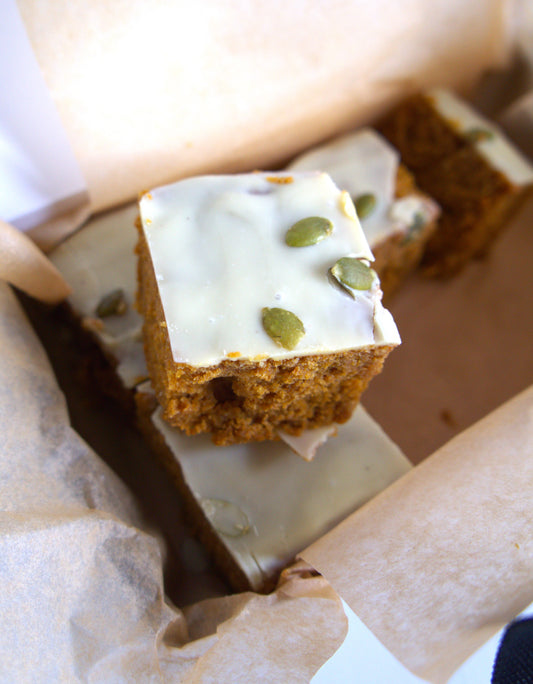 Square piece of blondie cake with white frosting and green pumpkinseeds in a box.